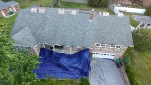 An aerial view of a house with a gray shingle roof, bundles of new shingles on the roof, and tarps below.
